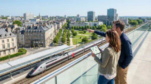 Couple sur un pont dominant une ville et une gare TGV. La femme regarde une tablette avec une carte; l'homme pointe le paysage.