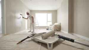 Un peintre en blanc applique de la peinture claire sur un mur. Au premier plan, un bac à peinture, un rouleau et un pinceau sur une bâche.